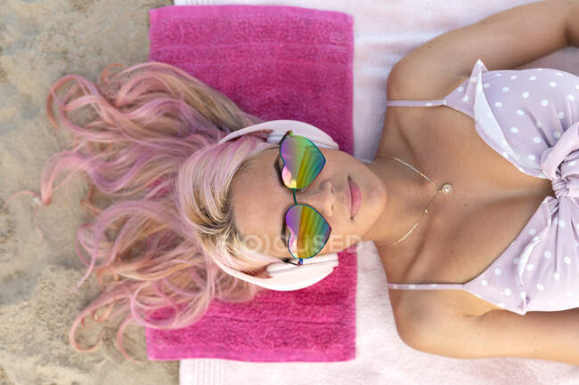Vista dall'alto della femmina con i capelli rosa e gli occhiali da sole sdraiati su un asciugamano sulla riva sabbiosa e rilassanti durante le vacanze estive ascoltando musica in cuffia — Foto stock