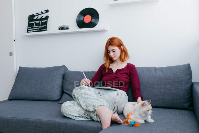 Giovane femmina a piedi nudi con capelli rossi navigazione internet su tablet mentre seduto con gatto sul divano a casa — Foto stock