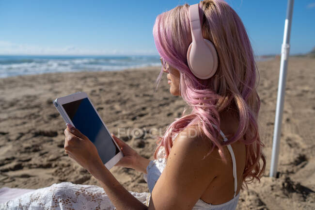 Side view of female with pink hair chilling on seashore while listening to music in headphones and surfing Internet on tablet in summer — Stock Photo