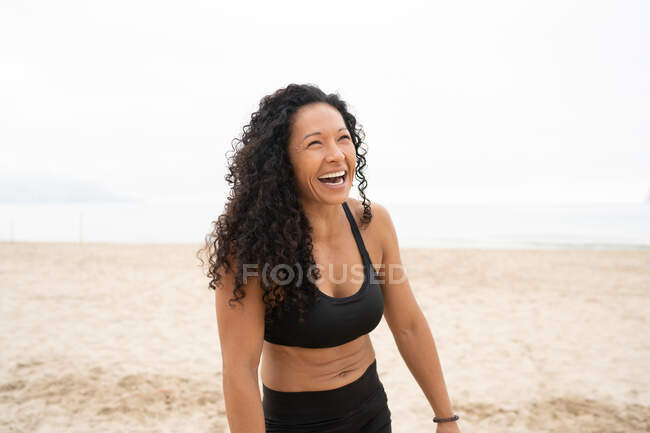 Позитивная азиатская спортсменка с вьющимися волосами, смеющаяся на песчаном побережье летом — стоковое фото