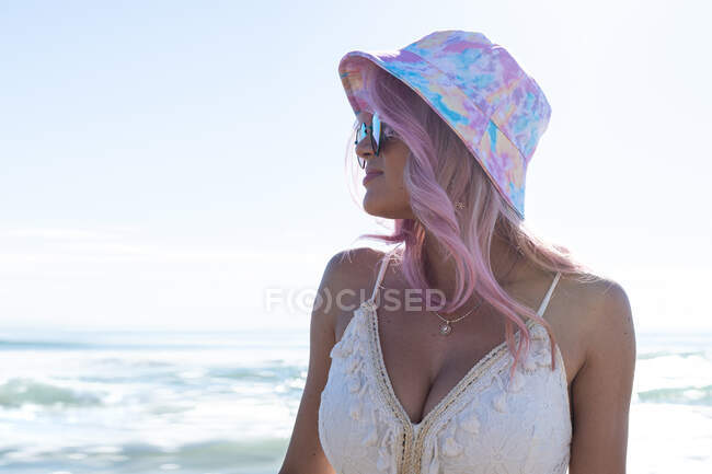 Femme contente avec des cheveux roses et en robe d'été profitant debout sur la plage contre la mer et regardant loin — Photo de stock