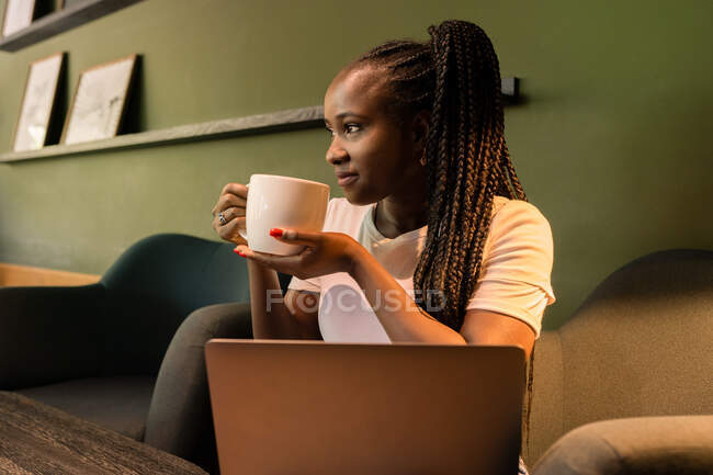 Encantada hembra afroamericana sentada en la cafetería con una taza de café y disfrutando del fin de semana mientras mira hacia otro lado - foto de stock