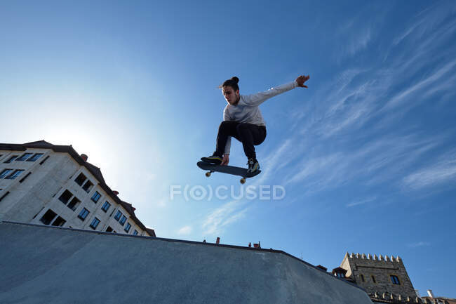 De baixo de jovem bravo patinador masculino pulando com skate e realizando acrobacias no parque de skate no dia ensolarado contra o céu azul — Fotografia de Stock