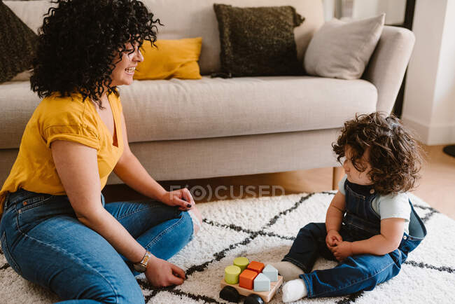 Mãe no desgaste casual brincando com a criança pequena bonito enquanto se sentam no chão juntos na sala de estar — Fotografia de Stock