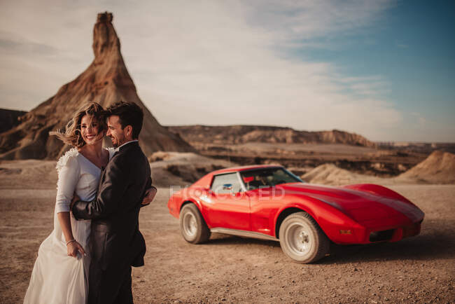 Side view of cheerful bride and groom embracing each other while standing near red luxury vehicle during road trip through Bardenas Reales Natural Park in Navarra, Spain — Stock Photo