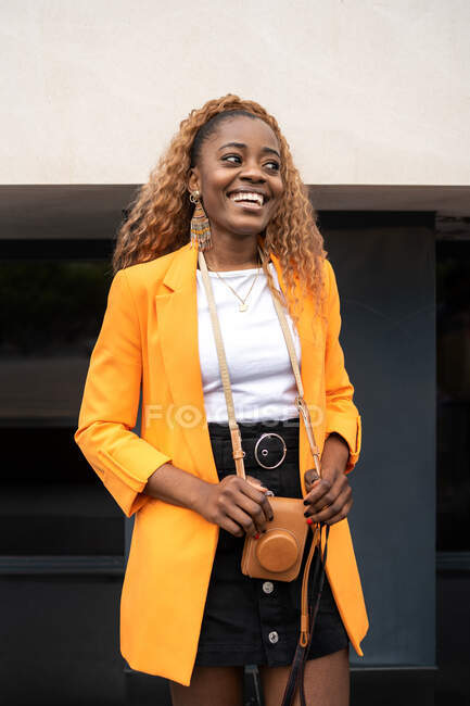 Femme afro-américaine positive aux cheveux bouclés souriant largement tout en se tenant debout avec un appareil photo et en regardant ailleurs — Photo de stock