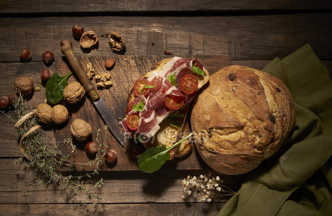 Top view of appetizing homemade sandwich made with freshly baked artisan bread with sliced prosciutto and tomatoes on rustic wooden table with nuts and herbs — Stock Photo