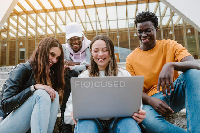 Diverse junge Freunde lächeln breit, während sie gemeinsam auf Betontreppen sitzen und Videos auf Netbook ansehen — Stockfoto