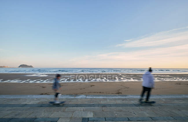 Junge aktive männliche Freunde fahren gemeinsam Skateboards entlang der Promenade vor dem Hintergrund von Meer und Sonnenuntergang — Stockfoto