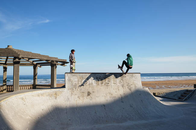 Patinadores activos montando monopatines y mostrando trucos en skate park en la playa en verano - foto de stock