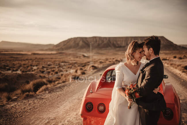 Happy bride and groom sorridente e abbracciarsi mentre si appoggia sul veicolo di lusso rosso durante il viaggio in auto attraverso il Parco Naturale Bardenas Reales in Navarra, Spagna — Foto stock