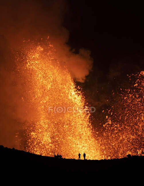Silhouettes of anonymous travelers standing against orange fume of active volcano in Iceland — Stock Photo