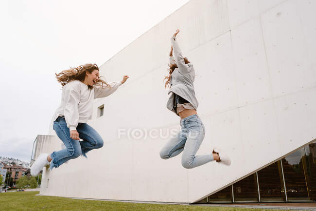 Low angle full body joyful young female best friends jumping together above ground while enjoying pleasant time — Stock Photo