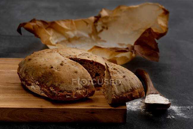 Композиція зі свіжоспеченим сільським кислим круглим хлібом на пергаментному папері, розміщеному на дерев'яній дошці з ложкою і пшеничним борошном — стокове фото