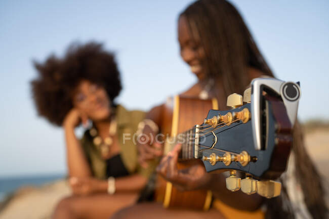 Happy young African American female friends playing guitar while sitting together on sandy seashore and enjoying summer holidays — Stock Photo