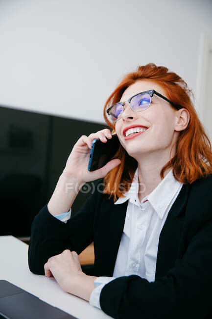Young female entrepreneur in formal wear talking on cellphone while working at desk with laptop at home — Stock Photo