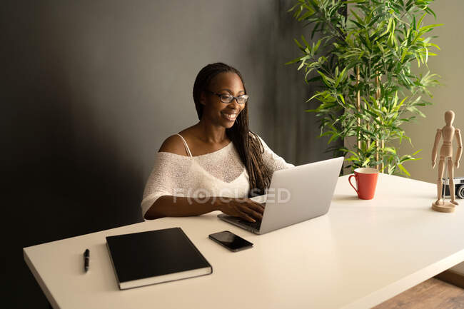 Fröhliche afroamerikanische Freelancerin sitzt am Tisch am modernen Arbeitsplatz und blättert im Netbook, während sie fernab von zu Hause an einem Projekt arbeitet — Stockfoto