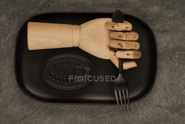 Mão de madeira artificial com garfo colocado na bandeja perto da lata preta selada com conservas na mesa — Fotografia de Stock