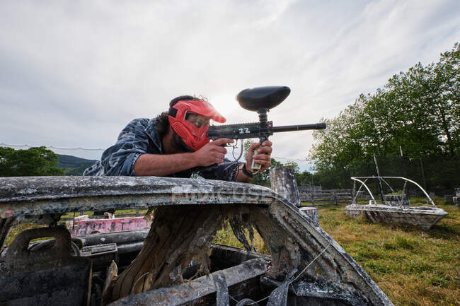 Jogador masculino em roupa de camuflagem e com arma deitada no carro abandonado e apontando enquanto joga paintball no campo — Fotografia de Stock