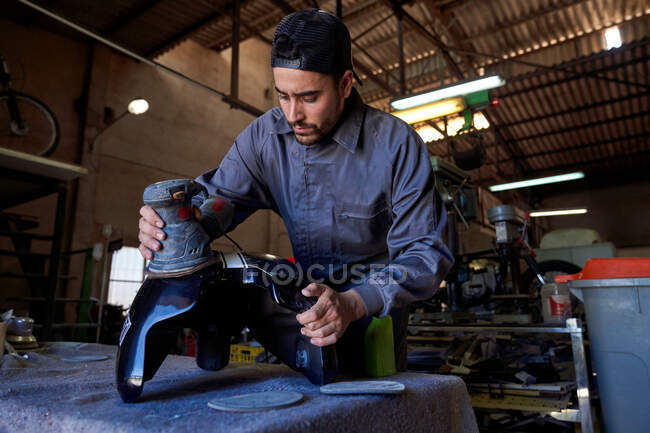 Low angle of concentrated male worker standing at workbench and polishing motorcycle tank with electric instrument while working in garage — Stock Photo