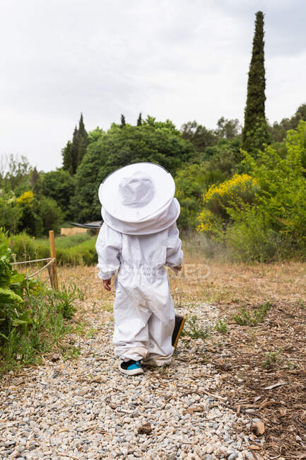 Back view of unrecognizable kid in white beekeeper protective costume standing near metal bee smoker in village — Stock Photo