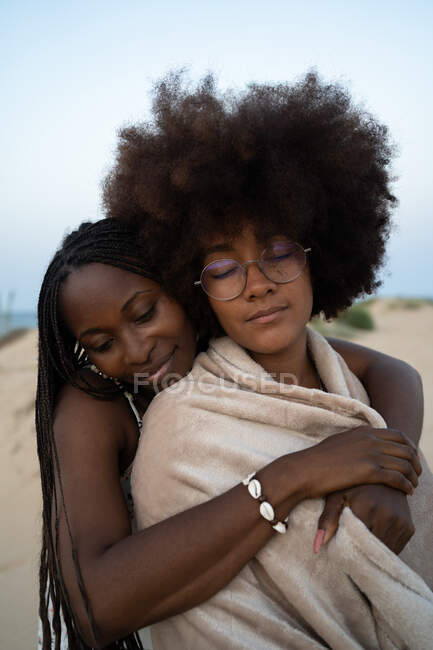 Happy young multiracial female best friends wrapped in warm blanket cuddling with eyes closed while chilling and enjoying time together in summer evening during holidays on seashore — Stock Photo