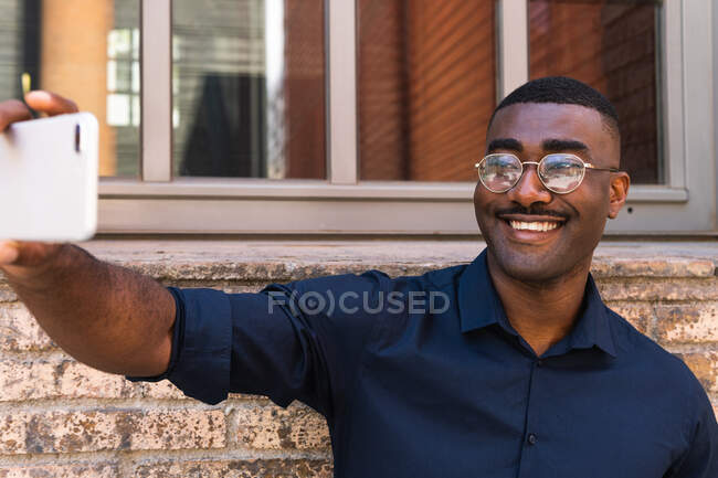 Heureux homme noir adulte en lunettes et chemise prendre selfie sur téléphone portable dans la rue de la ville en plein jour près de bâtiment en brique avec fenêtre — Photo de stock