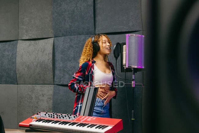 Contenu jeune chanteuse noire dans les écouteurs toucher le ventre tout en chantant dans le microphone contre synthétiseur en studio de musique — Photo de stock