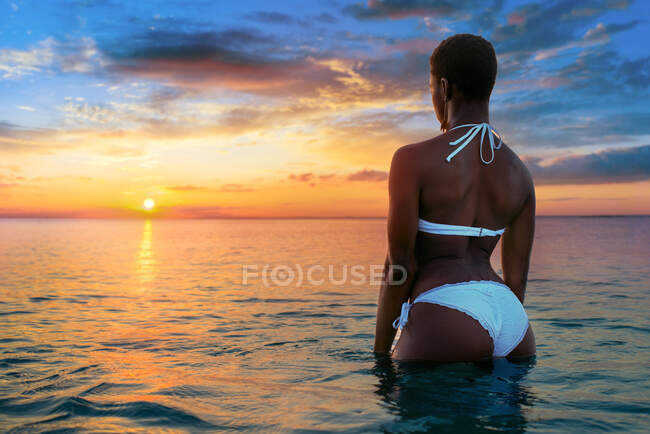 Visão traseira de uma mulher afro-americana irreconhecível de biquíni branco em pé na água do mar calma e admirando o incrível céu colorido ao pôr do sol no verão — Fotografia de Stock