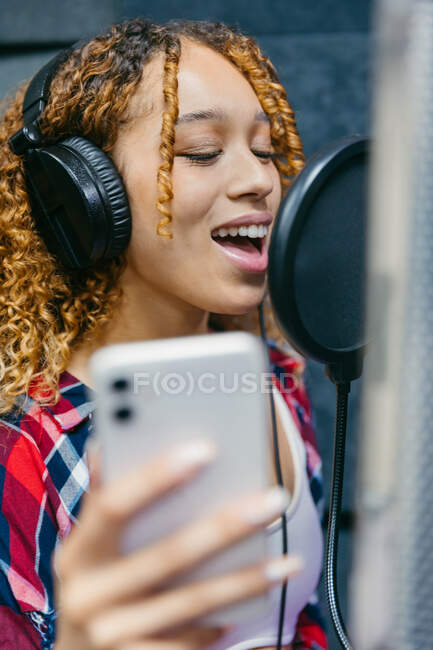 Cheerful African American female singer in headphones with smartphone performing song against sound shield in recording studio — Stock Photo