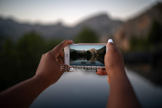 Cultivo irreconocible viajera afroamericana tomando fotos en el teléfono inteligente del estanque en las tierras altas - foto de stock