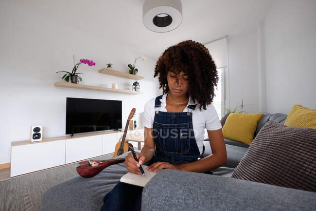 Thoughtful African American female musician sitting on bed with mandolin and notebook while composing song at home — Stock Photo