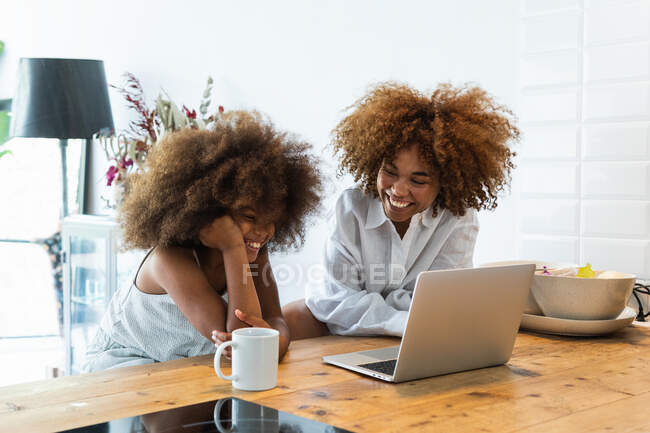 Afro-américaine femme et fille avec afro coiffure assis à la table de la cuisine et la navigation netbook ensemble le week-end matin à la maison — Photo de stock