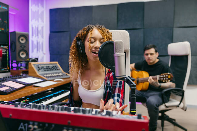 Guitarrista masculino feliz tocando guitarra acústica contra parceiro feminino preto em fones de ouvido cantando no microfone em estúdio de gravação — Fotografia de Stock