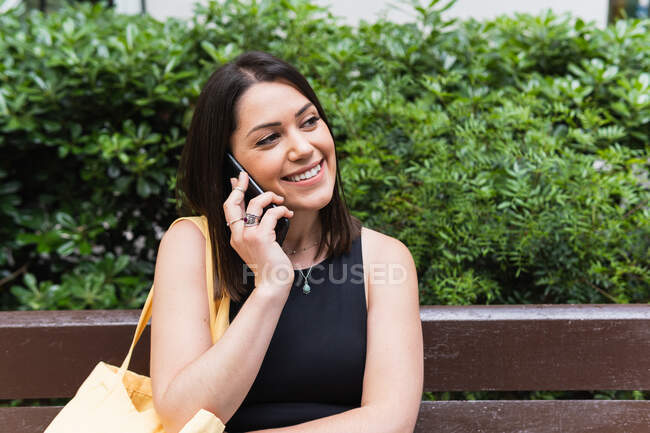Glückliche Frau spricht mit Handy, während sie auf Bank im Park sitzt und wegschaut — Stockfoto