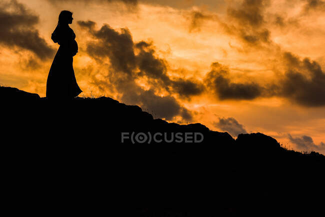 Low angle side view silhouette of anonymous pregnant female standing alone on hill and dreaming about future at sunset time — Stock Photo