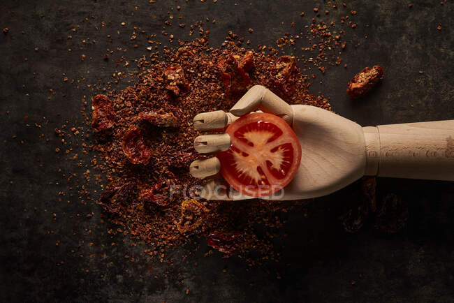 Top view composition with fresh red tomato slice in artificial wooden hand placed above ground sun dried tomatoes on black background — Stock Photo