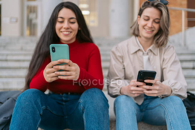 Two student girls sitting on the stairs watching the mobile — Stock Photo