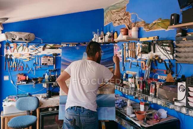 Visão traseira do artista masculino usando pistola de pulverização para pintar quadro em tela durante o trabalho em oficina criativa — Fotografia de Stock