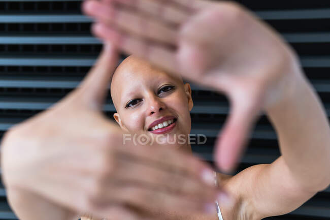 Encantada joven dama sin pelo con alopecia sonriendo mientras muestra el gesto de la cámara contra la pared jalousie en la calle - foto de stock
