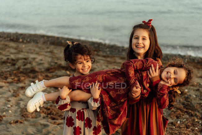 Симпатичні маленькі етнічні дівчата тримають маленьку сестру на руках, розважаючись разом на піщаному пляжі біля моря — стокове фото