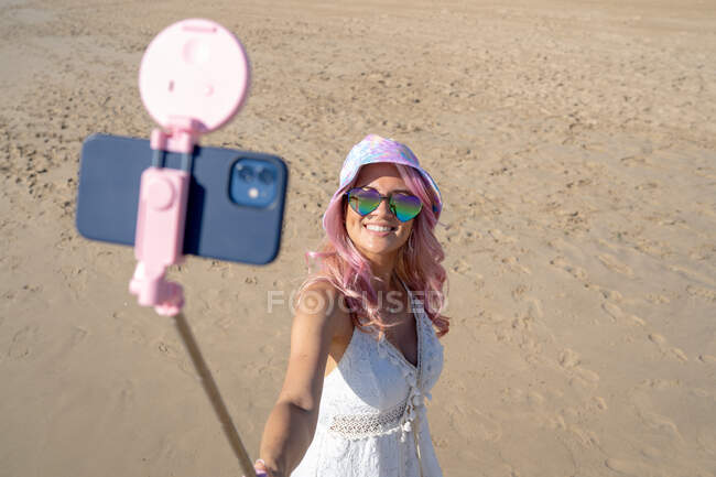 Высокий угол восторженной женщины с розовыми волосами и в летнем наряде делает самоснимок на смартфоне на селфи-палочке, стоя в солнечный день на берегу моря — стоковое фото