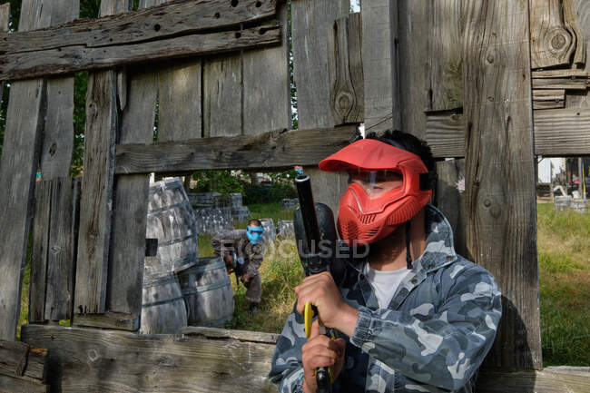 Jogador masculino em máscara e com pistola de paintball em pé atrás de cerca de madeira durante o jogo — Fotografia de Stock