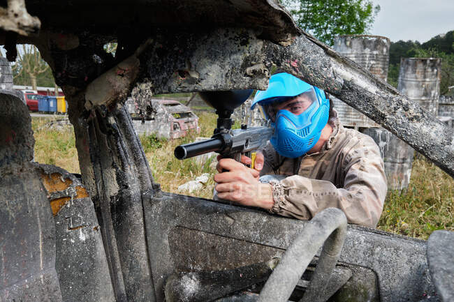 Мужчина в шлеме и с пистолетом играет в пейнтбол и прячется за заброшенной машиной во время игры — стоковое фото
