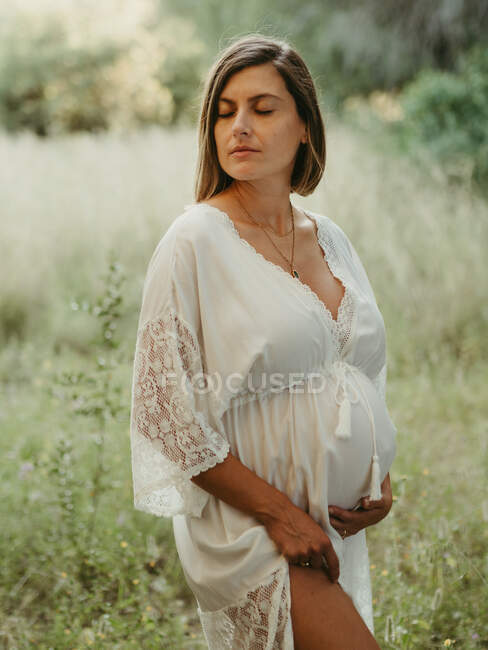 Спокойная беременная женщина в платье касается живота с закрытыми глазами, стоя в поле в сельской местности в летний день — стоковое фото
