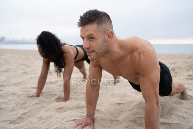Atletas deportivos multiétnicos masculinos y femeninos haciendo flexiones durante el entrenamiento físico en la orilla del mar de arena - foto de stock