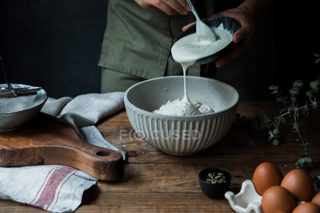 Неузнаваемый человек наливает йогурт в миску с мукой рядом с яйцами и семенами во время приготовления теста на деревянном столе — стоковое фото