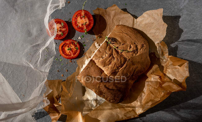 Сверху свежий ароматный хлеб ручной хлеб буханка на пергаментной бумаге помещен рядом нарезанные спелые красные помидоры и травы — стоковое фото