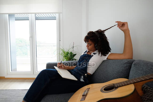 Спокойная афроамериканка-музыкантка, сидящая на кровати с гитарой и ноутбуком, сочиняя дома песню — стоковое фото