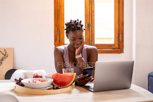 Entzückte Afroamerikanerin surft am Handy, während sie zu Hause am Tisch mit reifen, gesunden Früchten sitzt — Stockfoto